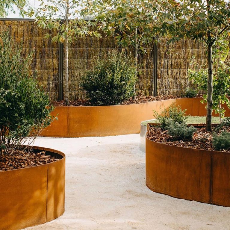 Curved rust-colored metal planters filled with shrubs surround a pale gravel path in a landscaped garden.