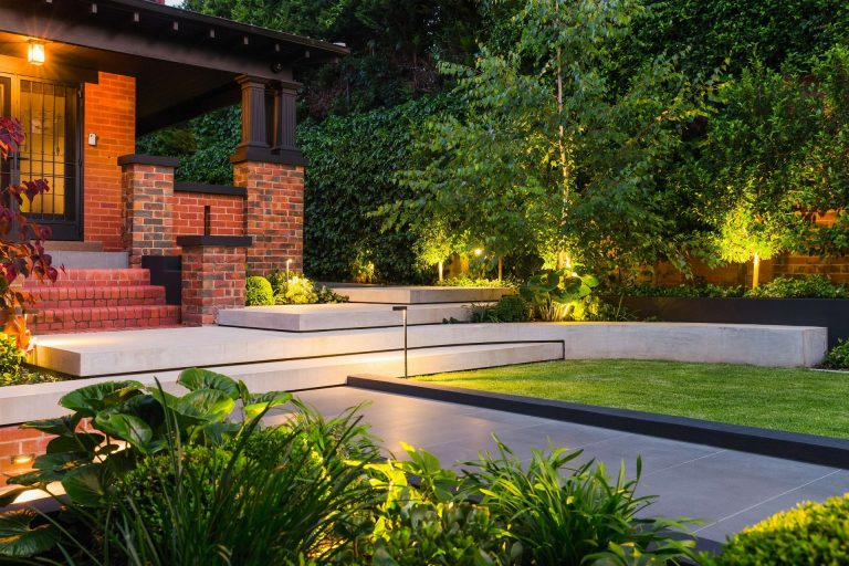 Brick house entry with stepped concrete terraces, warm lights, and dense greenery at dusk.